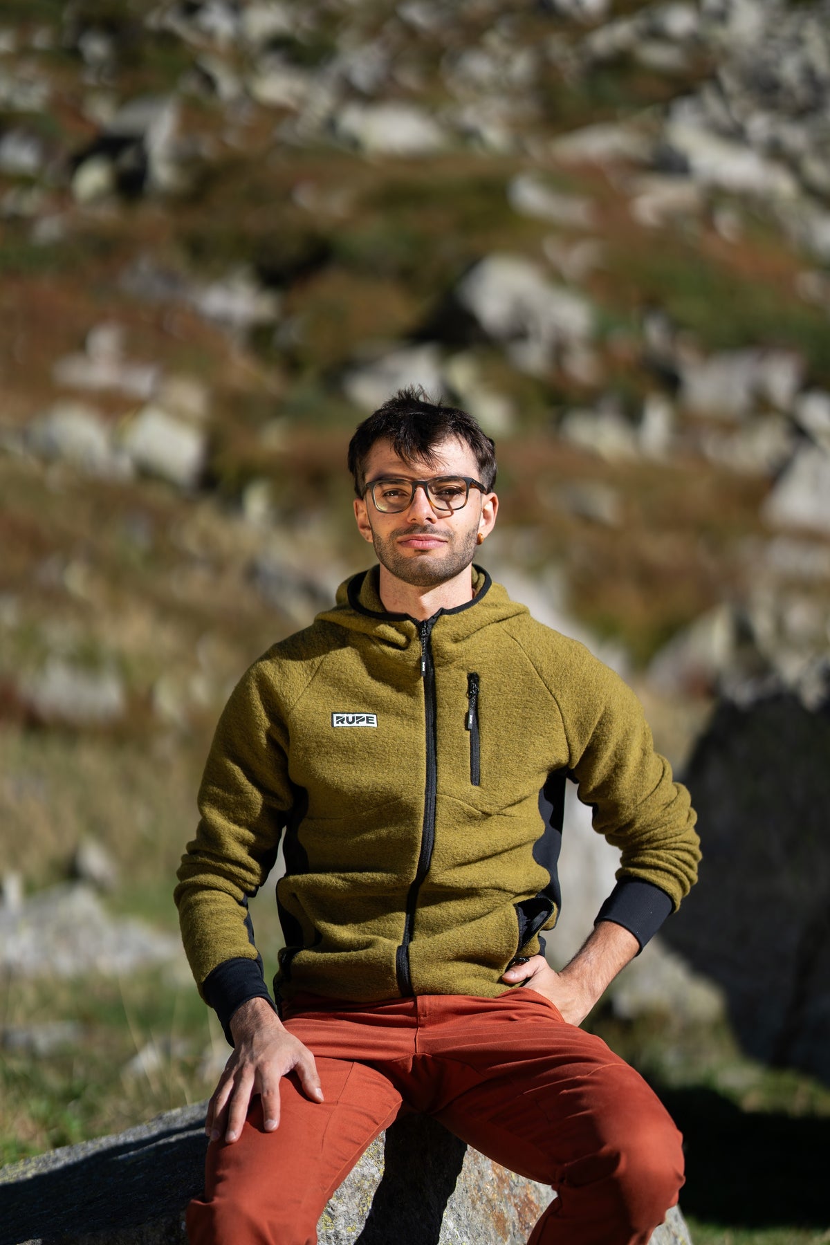 FW26-076 Summit Olive + Pirenei Rust (uomo seduto sul masso, frontale)